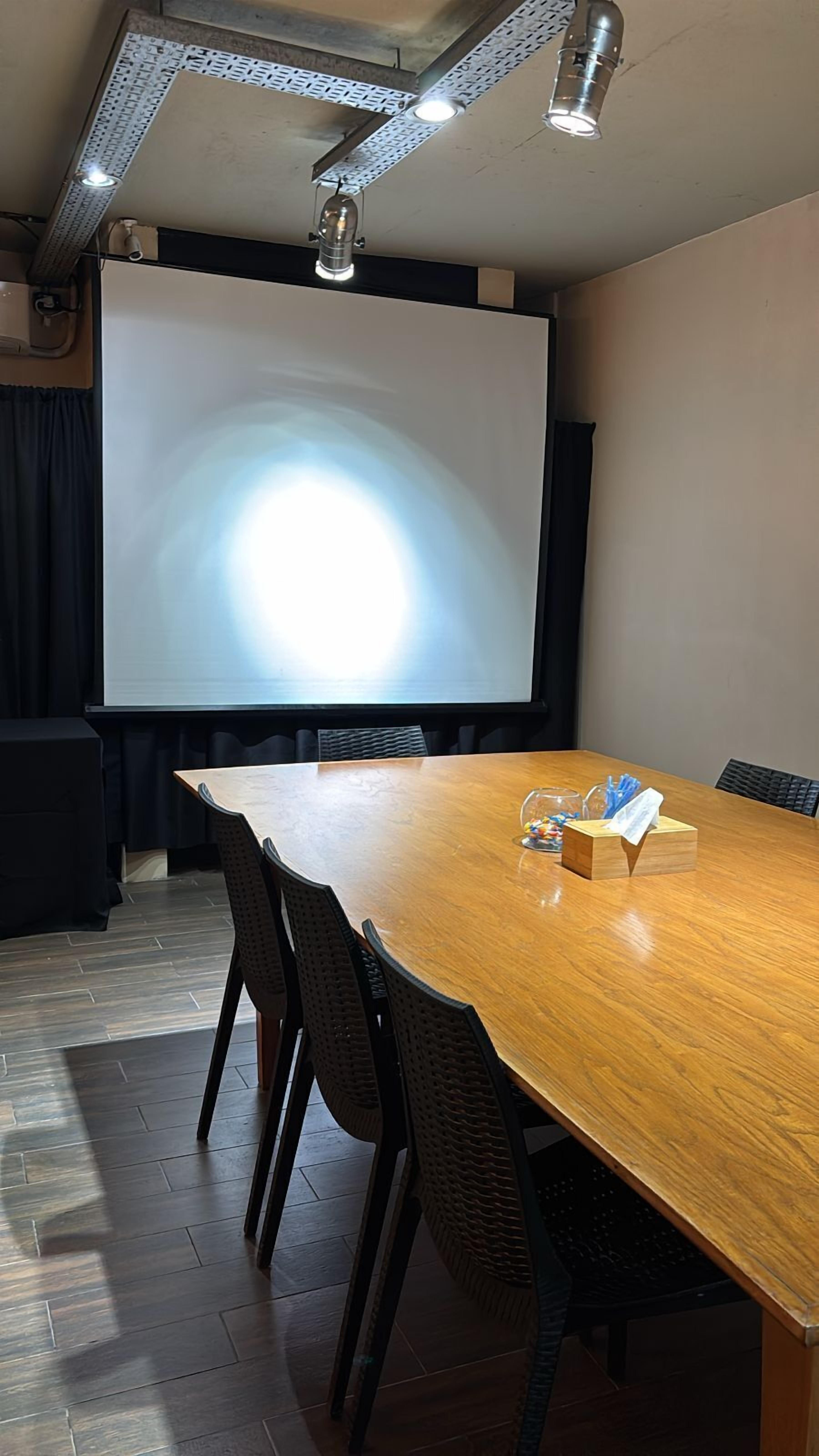 Een vergaderzaal met een lange houten tafel, zwarte stoelen, een projectiescherm en een whiteboard.