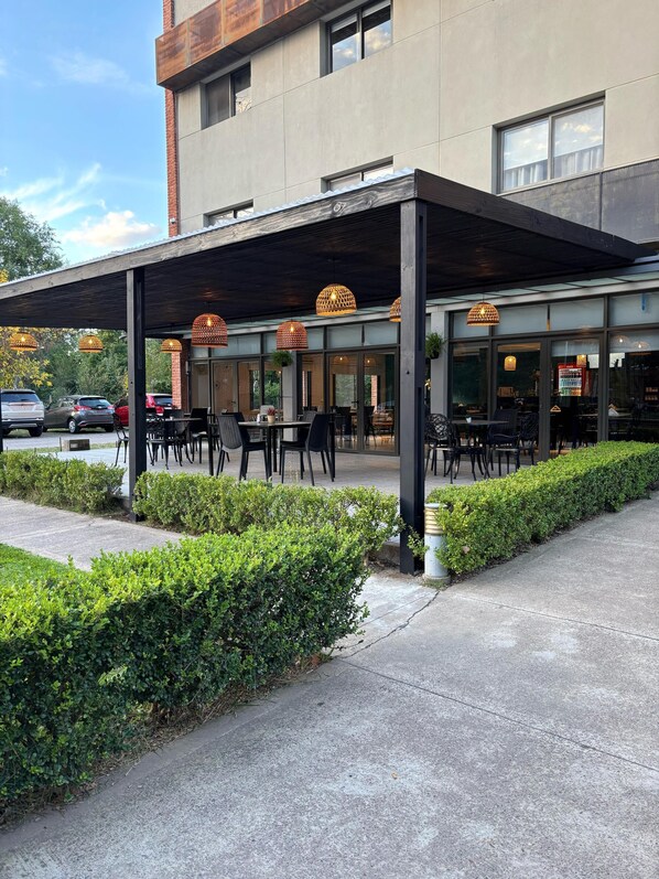 An outdoor seating area with tables and chairs under a covered patio, adjacent to a building with large windows. - Viva Suites Pilar (La Lonja)