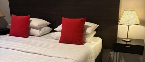 A hotel room with a large bed, two red pillows, a white bedspread, a framed picture on the wall, a nightstand with a lamp, and a small table.
