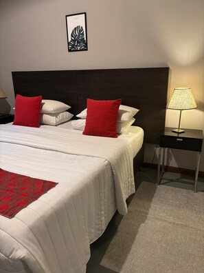 A hotel room with a large bed, two red pillows, a white bedspread, a framed picture on the wall, a nightstand with a lamp, and a small table. - Viva Suites Pilar (La Lonja)