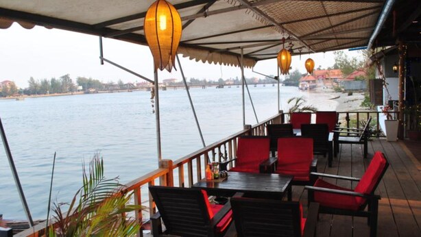 A restaurant with outdoor seating overlooking a body of water, featuring tables and chairs under a covered area with hanging lanterns. - The River Lodge (Kampot)