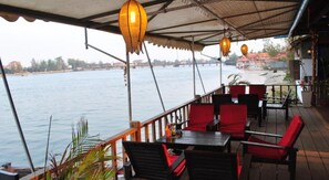 Ein Restaurant mit Terrassensitzgelegenheiten mit Blick auf ein Gewässer, ausgestattet mit Tischen und Stühlen unter einem Vordach mit hängenden Laternen.