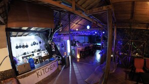 A kitchen area with a counter labeled "KITCHEN," a hanging pot rack, and a bar area with blue lighting. - The River Lodge (Kampot)