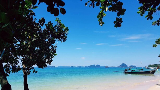A beach with clear turquoise water, a boat anchored near the shore, and a tree in the foreground. - Isle Beach Resort (Krabi)