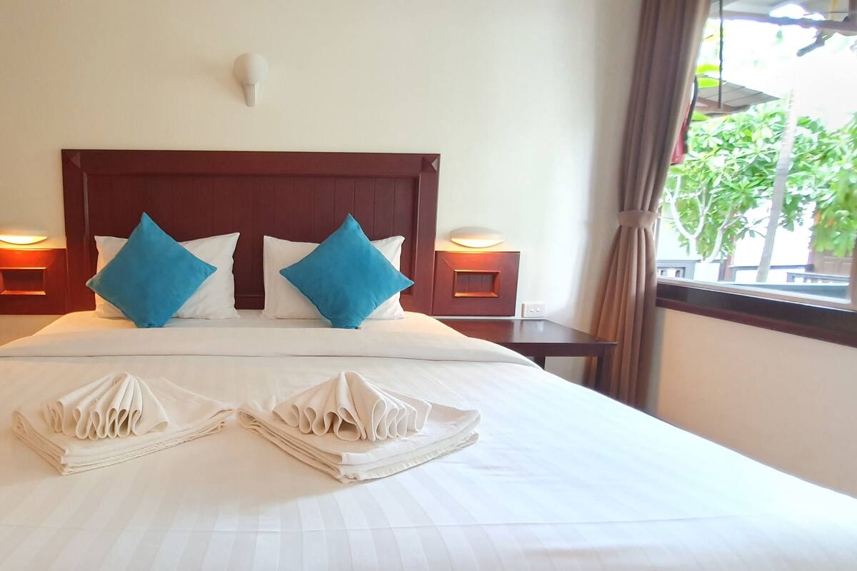 Quarto de hotel com uma cama grande, duas mesas de cabeceira com abajures e vista para o verde lá fora.