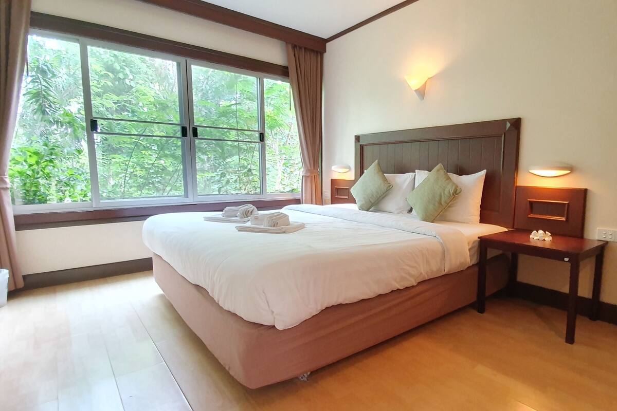 Cama bem arrumada com almofadas e uma mesa de cabeceira com abajur, janela com cortinas e vista para o verde lá fora.