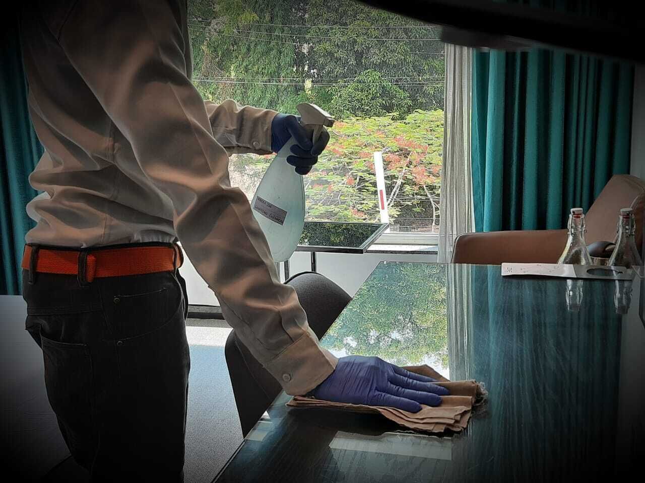 En person som torkar av ett bord med en sprayflaska, ett fönster med utsikt över grönska och ett bord med en flaska och glas.