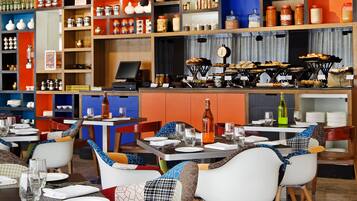 Un interior de restaurante con varias mesas y sillas, una caja registradora y estantes llenos de frascos y botellas.