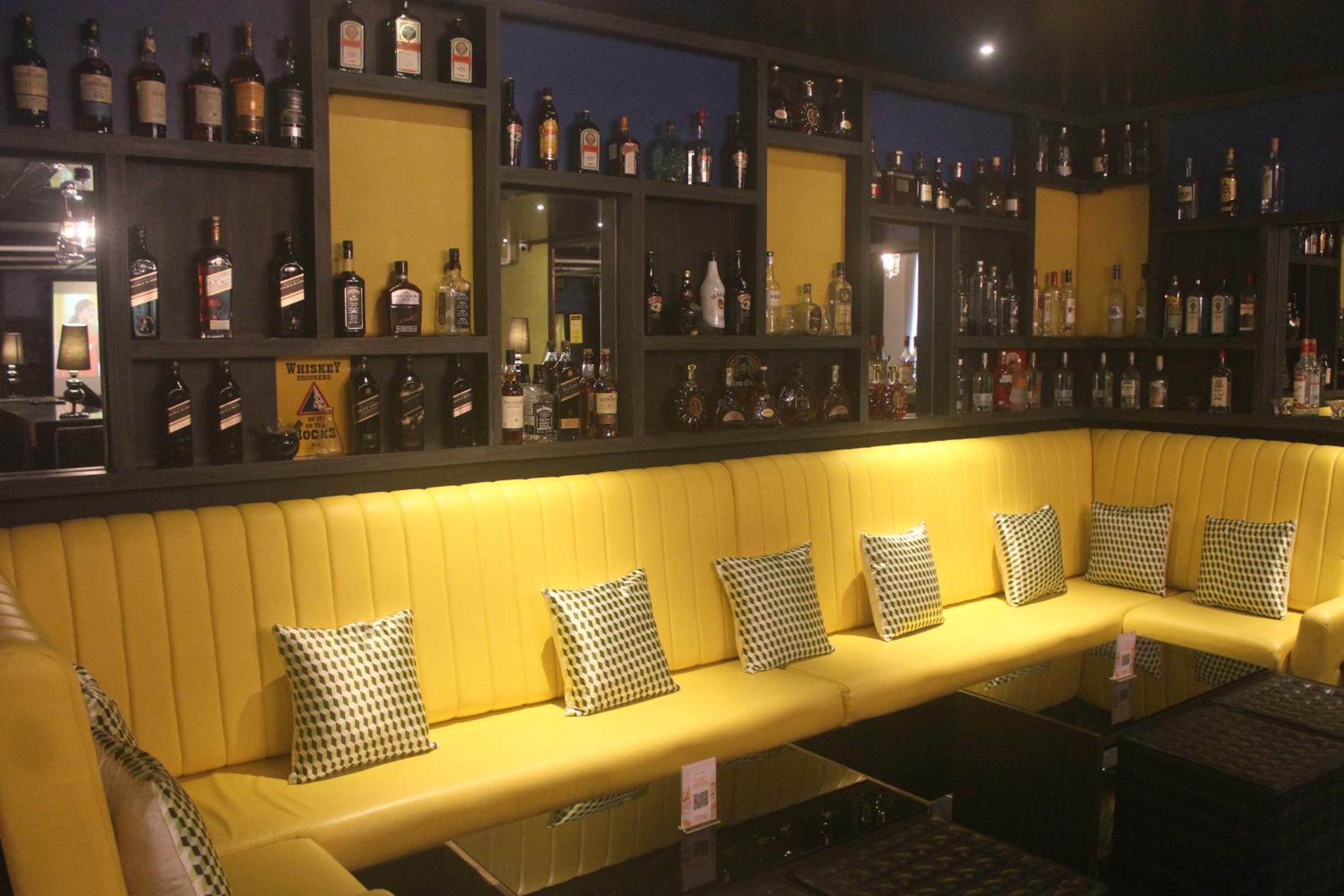 A bar area with a yellow cushioned bench, checkered pillows, and shelves of liquor bottles.