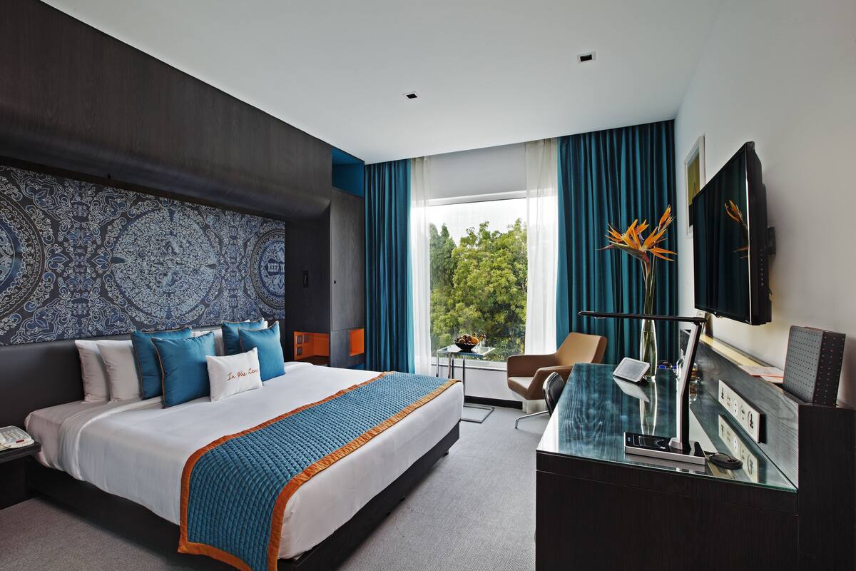A modern hotel room with a large bed, a desk with a computer, a TV, and a view of greenery outside.
