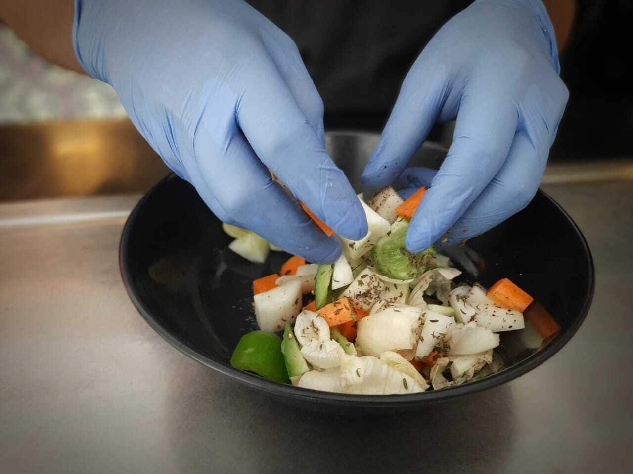Una persona con guantes azules preparando un plato con verduras en un tazón negro.