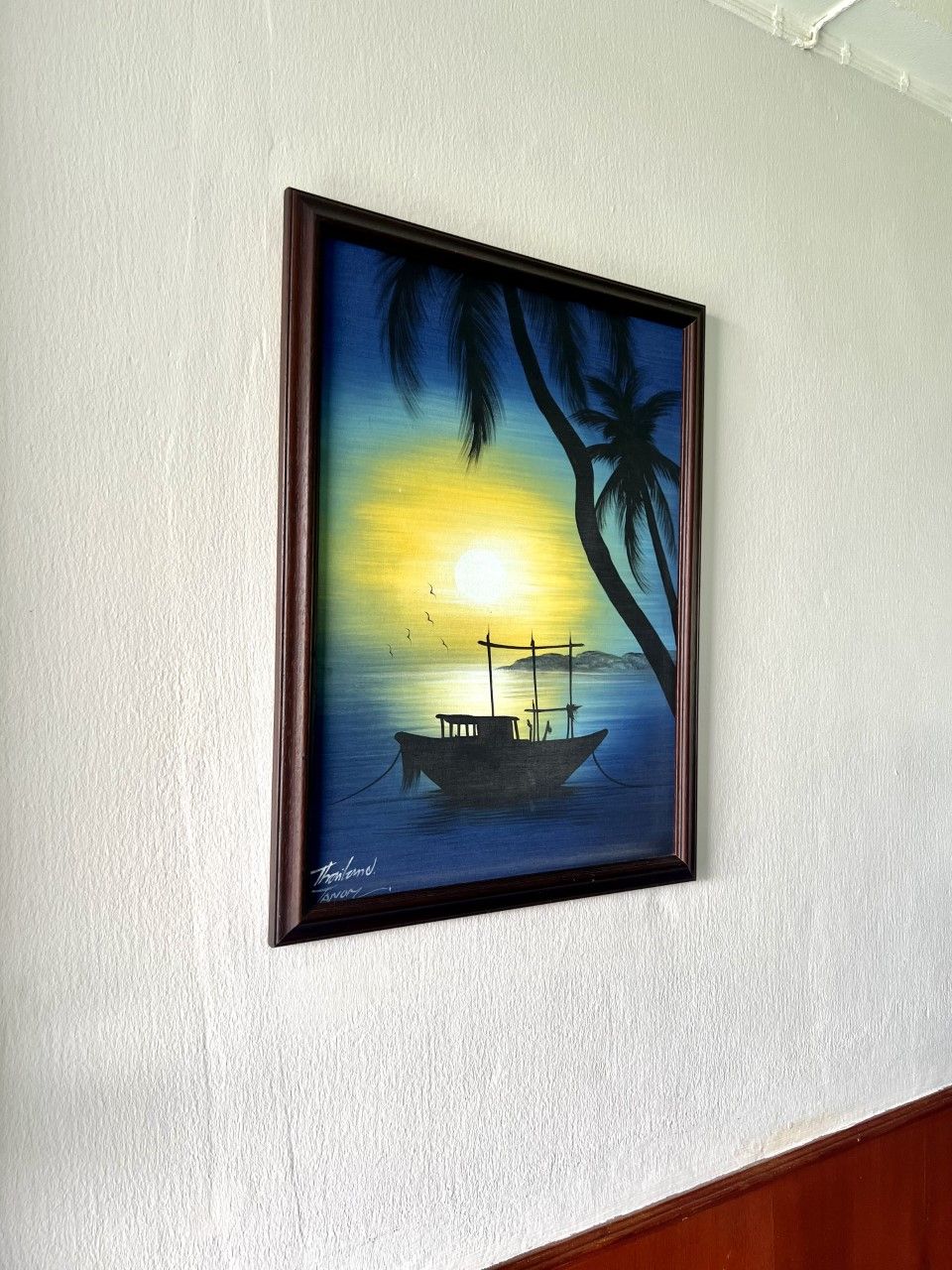 A framed painting of a sunset over water with a boat and palm trees on a white wall.