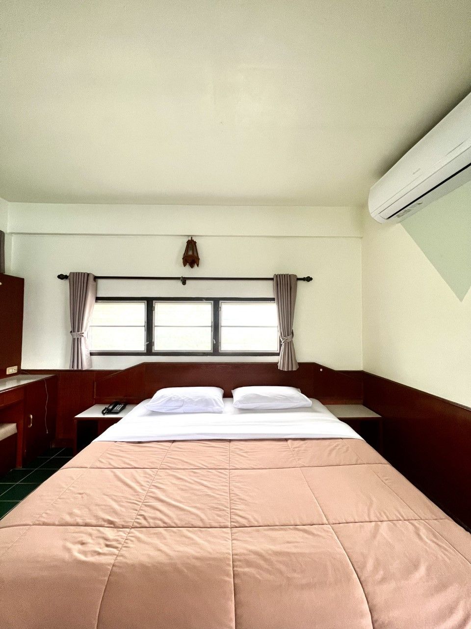A hotel room with a bed, a desk, a chair, a window with curtains, and an air conditioning unit.