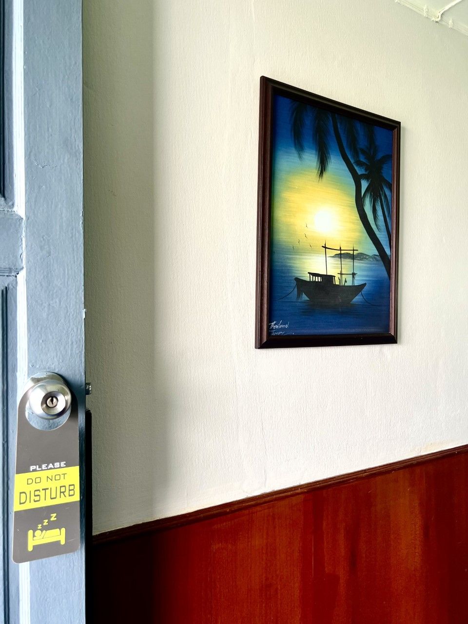 Una puerta con un letrero que dice 'PLEASE DO NOT DISTURB' y un cuadro enmarcado que muestra un barco en el agua con palmeras.