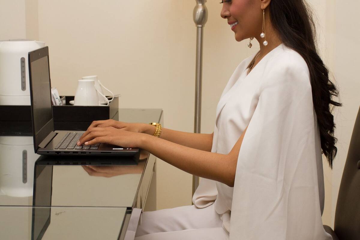A person seated at a desk with a laptop, a coffee maker, and a lamp in the background.