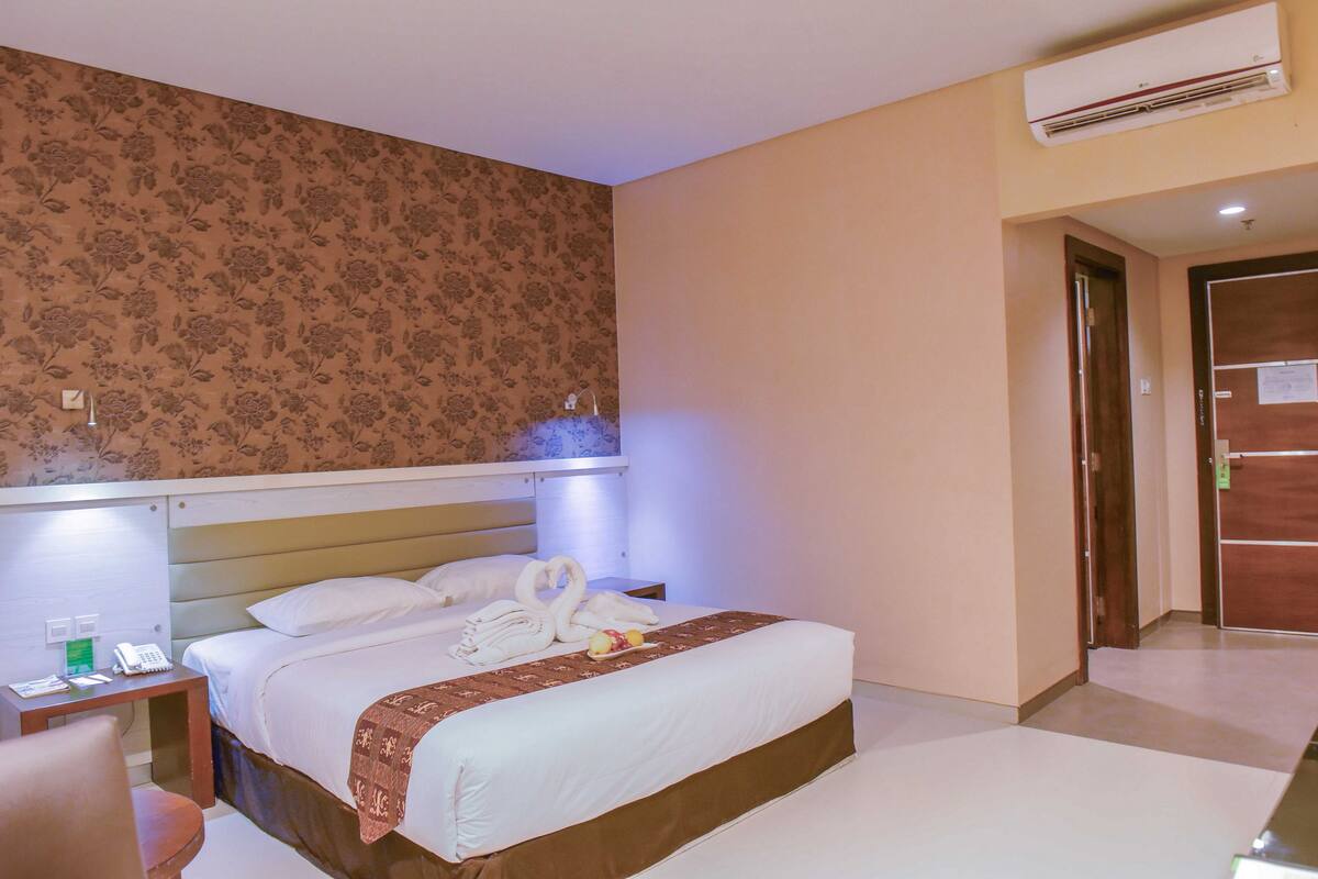 A hotel room with a bed, a bedside table, a chair, a desk, a telephone, and an air conditioning unit.