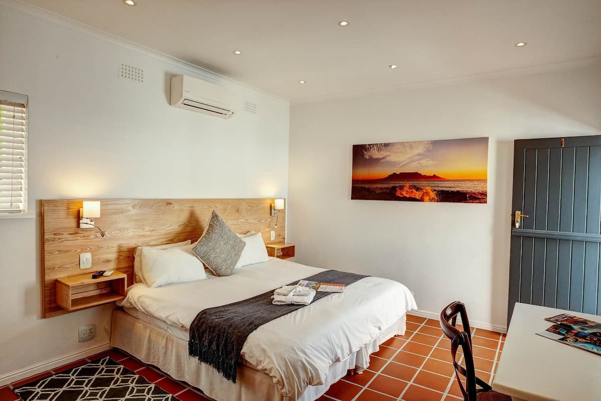 Ein Schlafzimmer mit einem Holz-Kopfteil, einem Bett mit weißer Bettwäsche, einem Nachttisch, einer Wand-Klimaanlage, einem Sonnenuntergangsgemälde, einer Tür und einem gefliesten Boden.