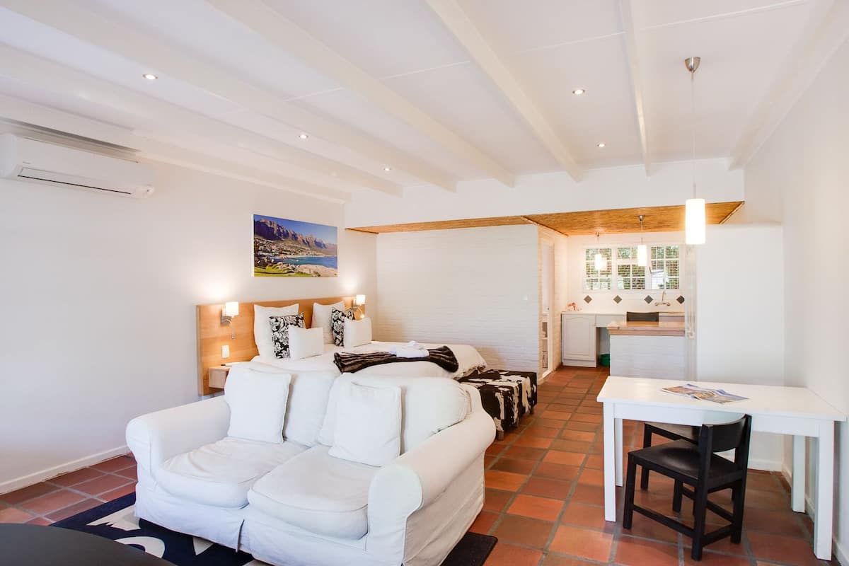 Ein moderner, gut beleuchteter Raum mit einem weißen Sofa, einem Bett mit gemusterten Kissen, einem kleinen Esstisch und einer Küchenzeile mit Fenster.
