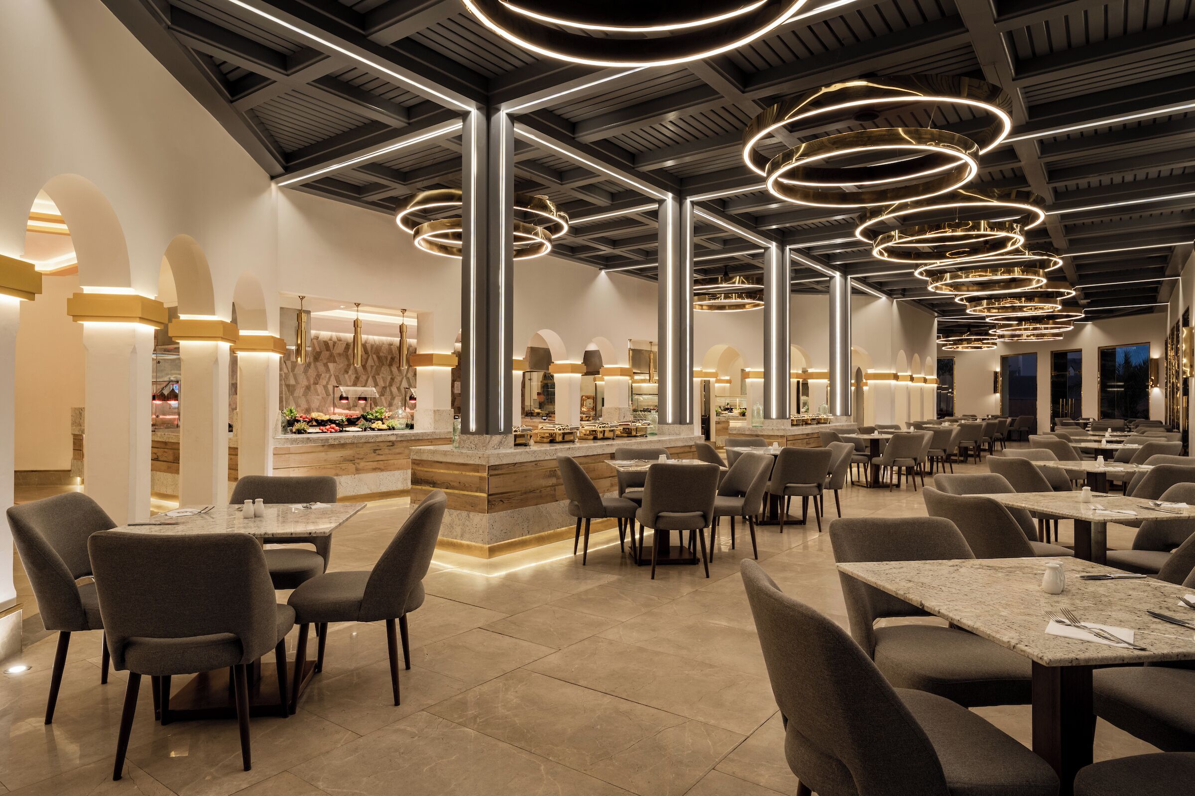 A spacious restaurant with multiple tables and chairs, a central bar area, and modern circular ceiling lights.
