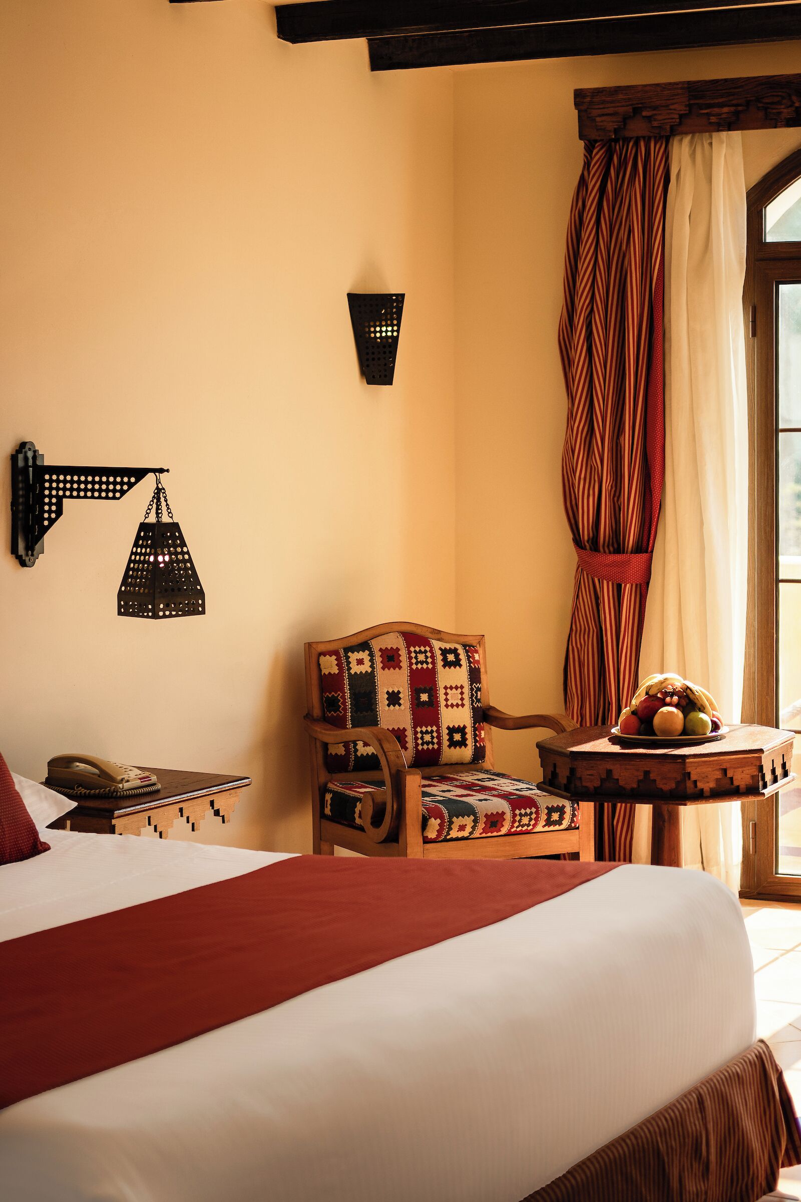 Habitación de hotel con una cama, una silla de madera con colcha, una mesita con fruta y una lámpara de pared.