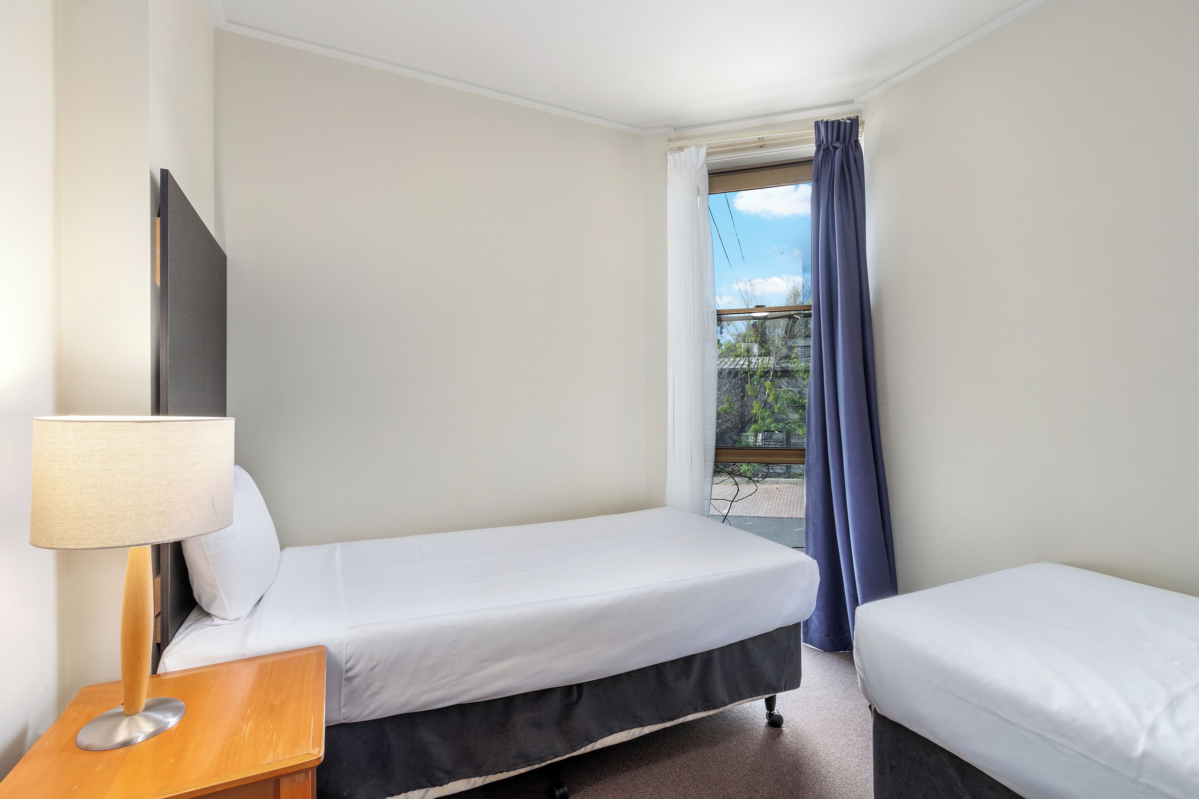 A hotel room with two single beds, a wooden nightstand with a lamp, and a large window with curtains.