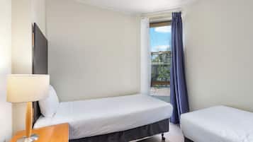 A hotel room with two single beds, a wooden nightstand with a lamp, and a large window with curtains.