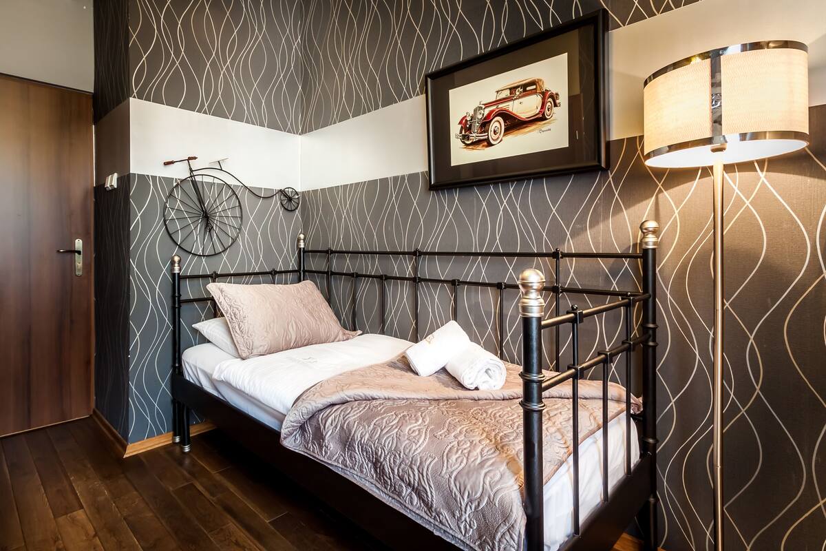 A neatly made bed with a headboard, a wall-mounted bicycle, a framed picture of a vintage car, and a floor lamp.