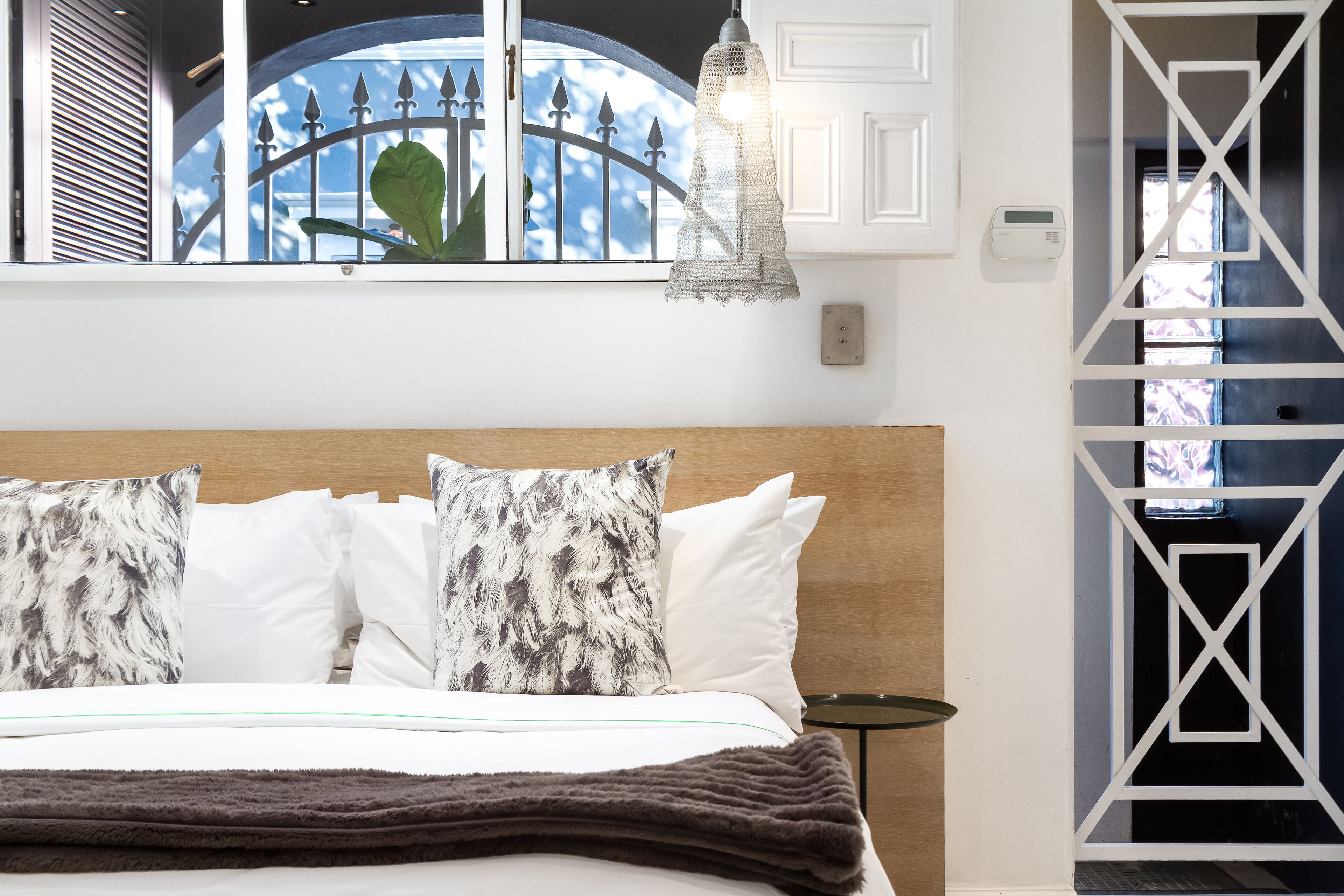 Una habitación de hotel con una cama, un ventanal con vistas a una valla y árboles, una lámpara colgante y un termostato en la pared.