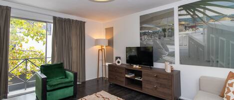 Una sala de estar moderna con un sillón verde, un televisor sobre un mueble de madera y un ventanal que da a un balcón.