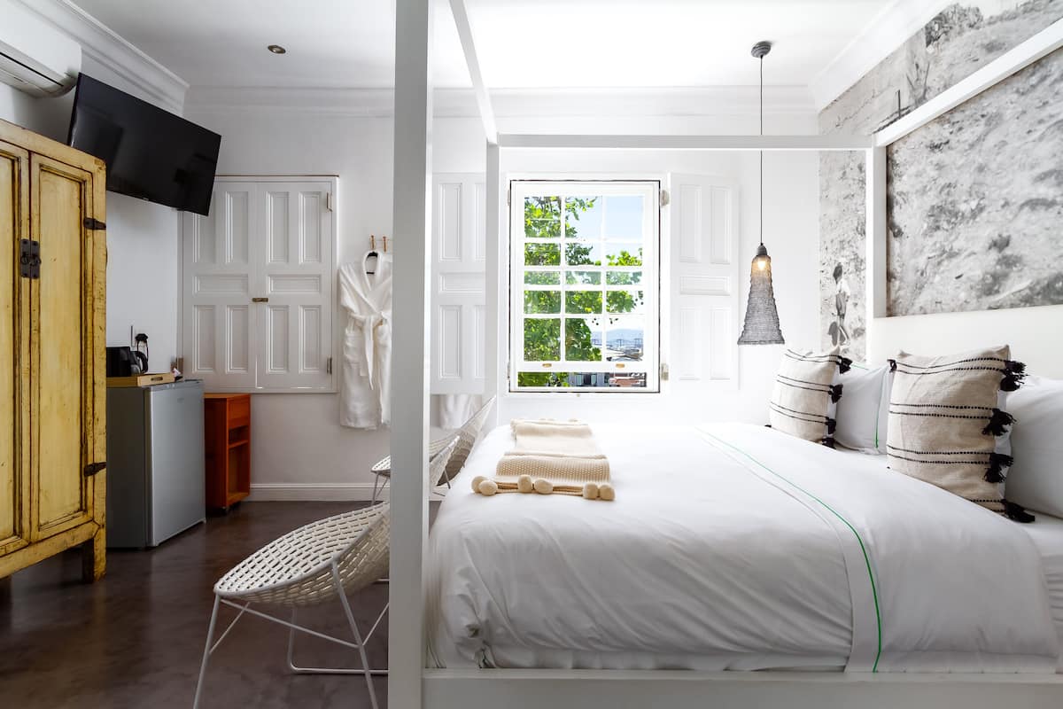 Quarto com cama, televisão, guarda-roupa e janela com vista para vegetação e um edifício.