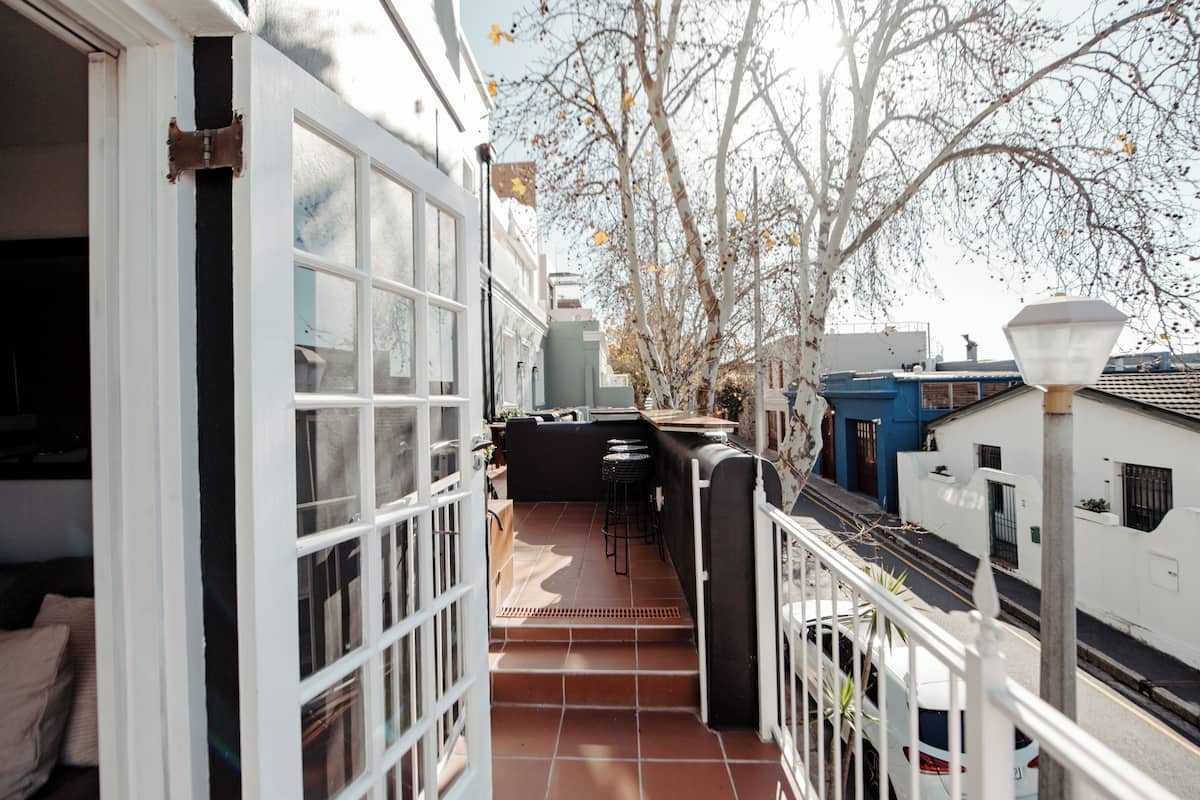 Um corredor exterior estreito com chão de azulejos, grades brancas e vista para a rua com árvores e edifícios.