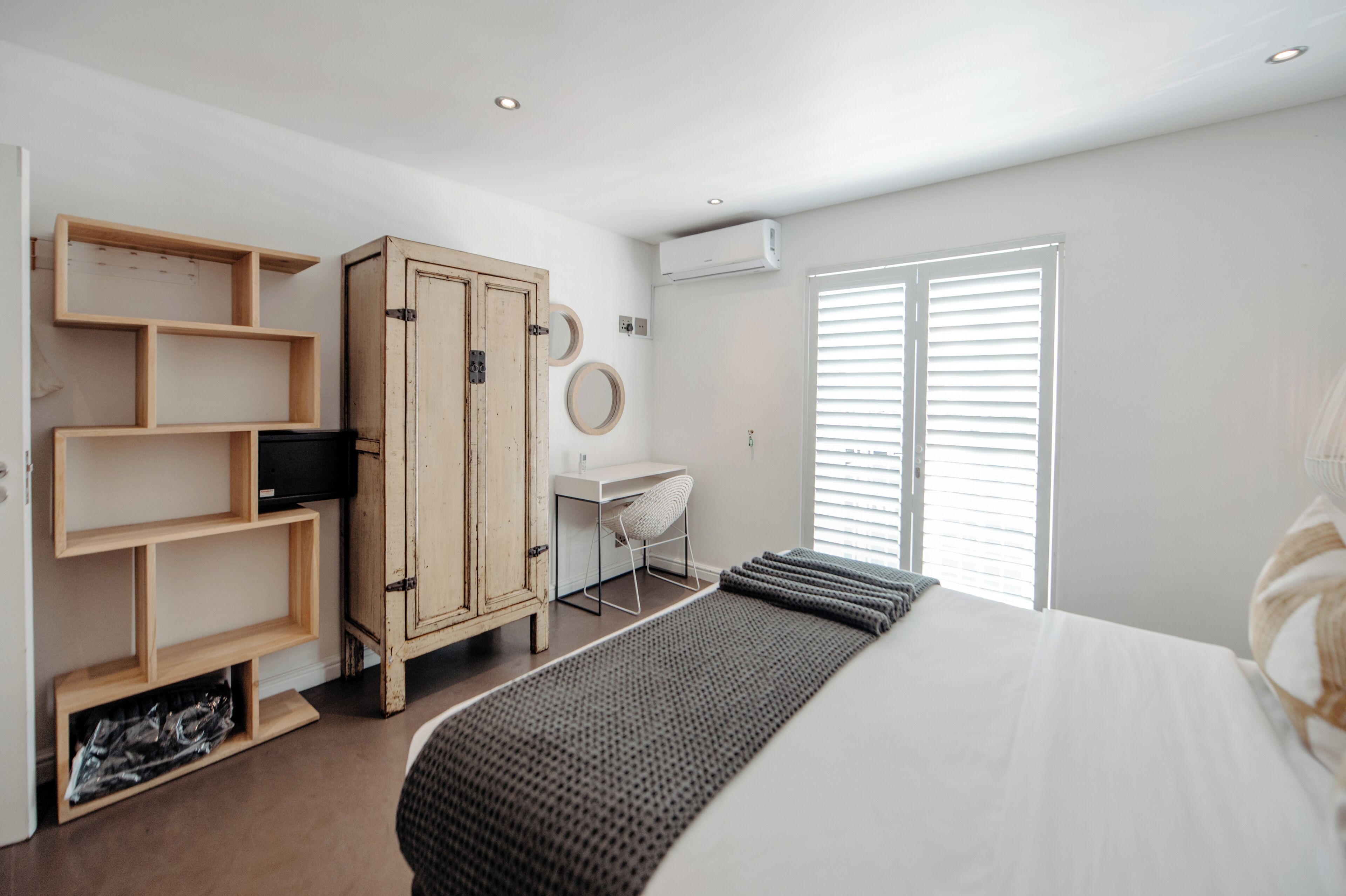 A bedroom with a bed, a wooden wardrobe, a desk, and a window with blinds.