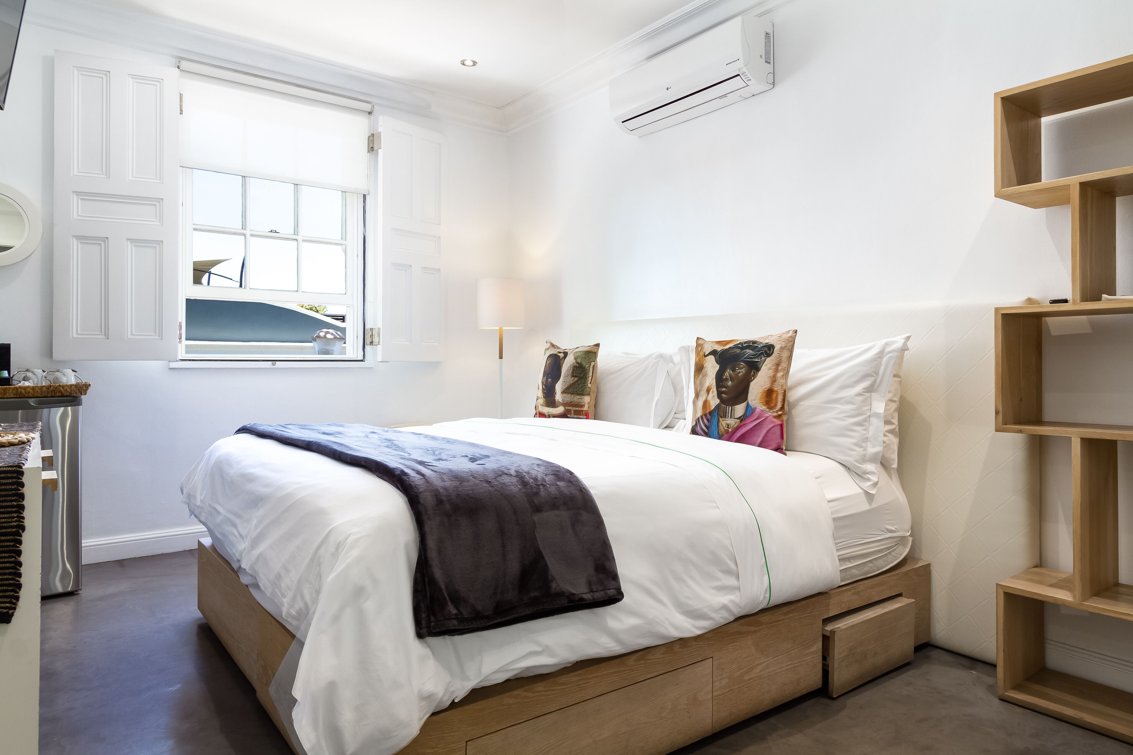 A bedroom with a bed, a window, a wall-mounted air conditioner, and a wooden shelf.