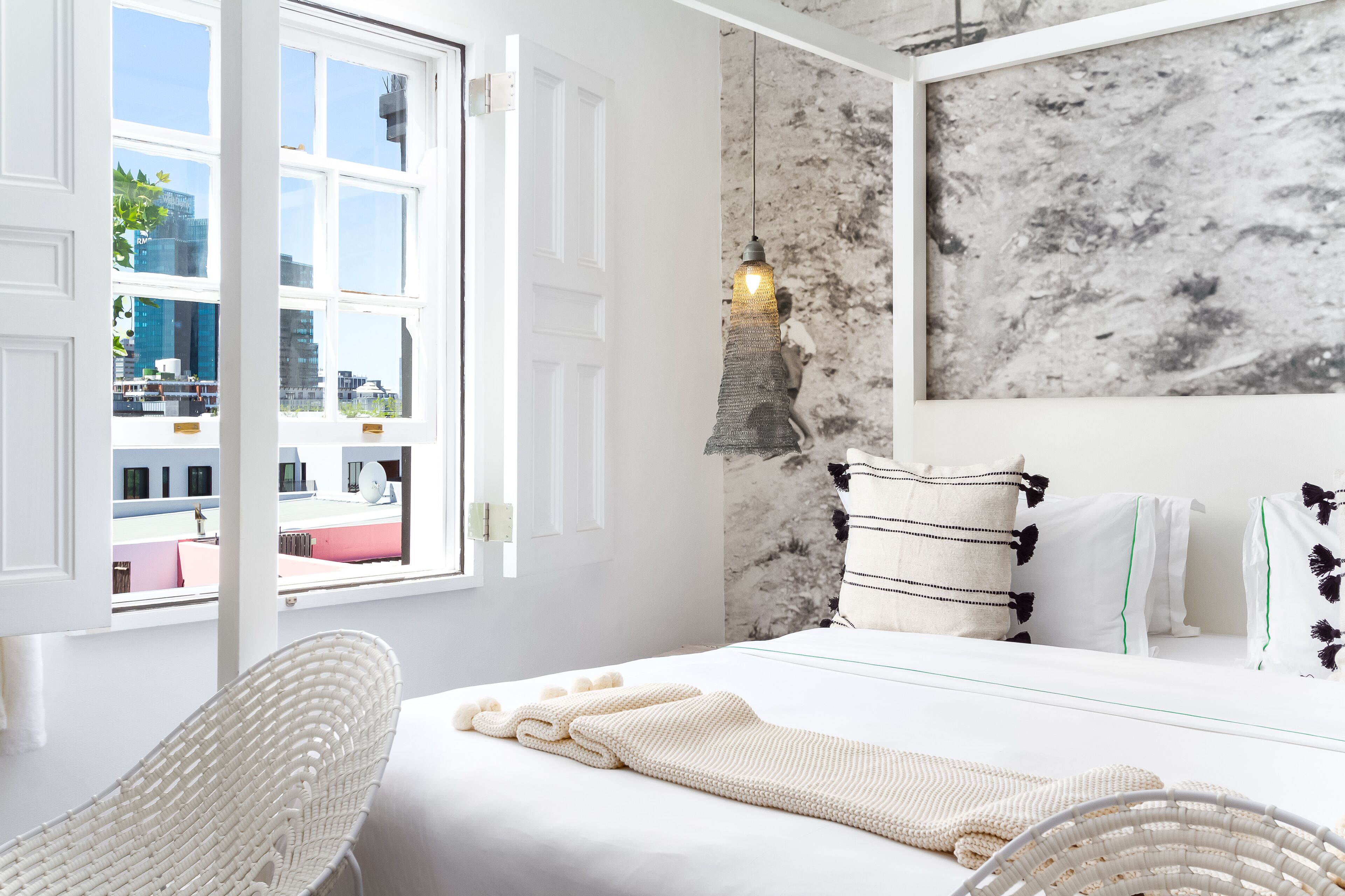 A modern hotel room with a large bed, a white chair, a view of the city through a large window, and a marble wall.