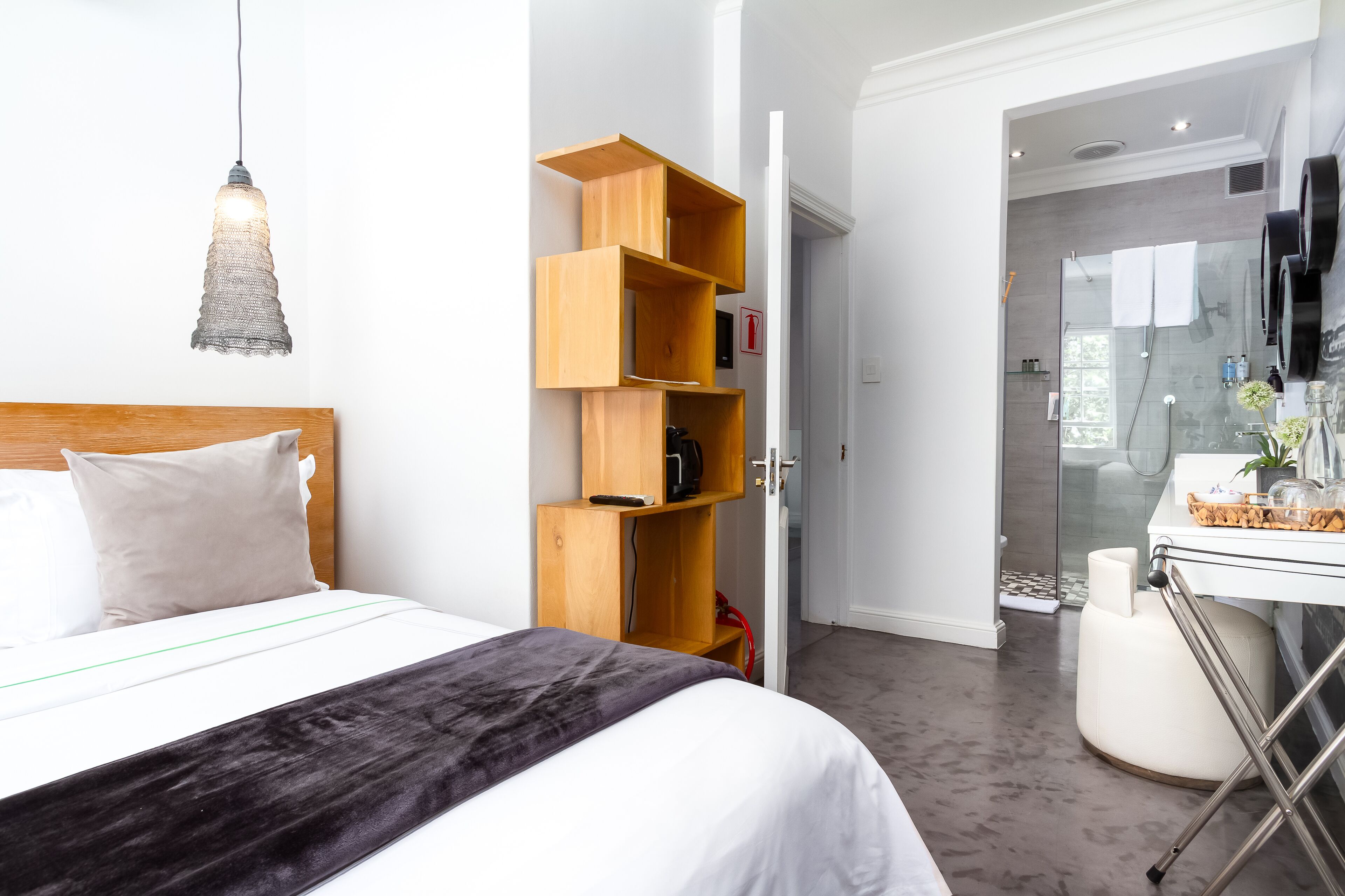 A modern hotel room with a neatly made bed, a wooden shelving unit, and a glass shower area.