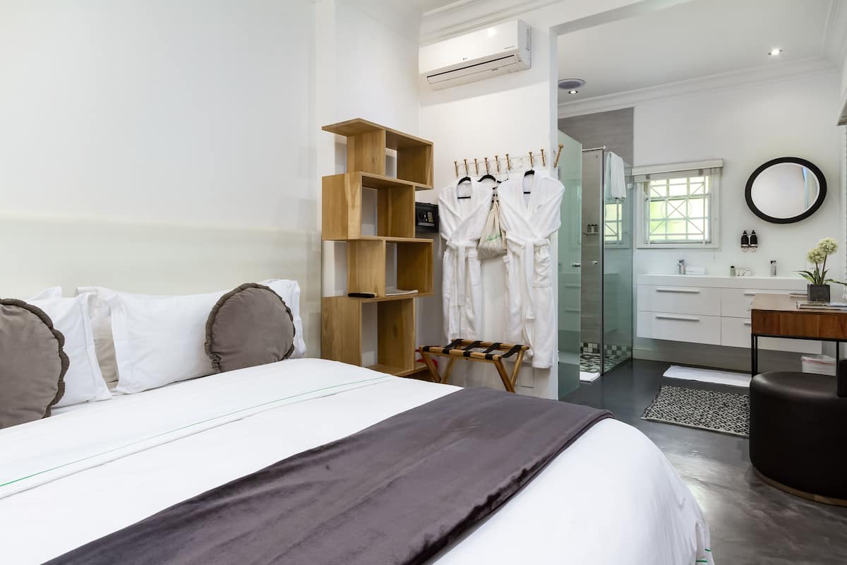 Quarto de hotel moderno com uma cama grande, uma estante de madeira, cabine de chuveiro de vidro, um espelho redondo e uma pequena área de estar.