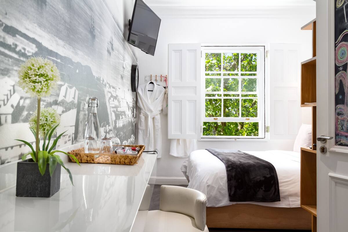 Quarto moderno com cama, televisão na parede, janela com vista para área verde e pequena mesa com planta e objetos decorativos.