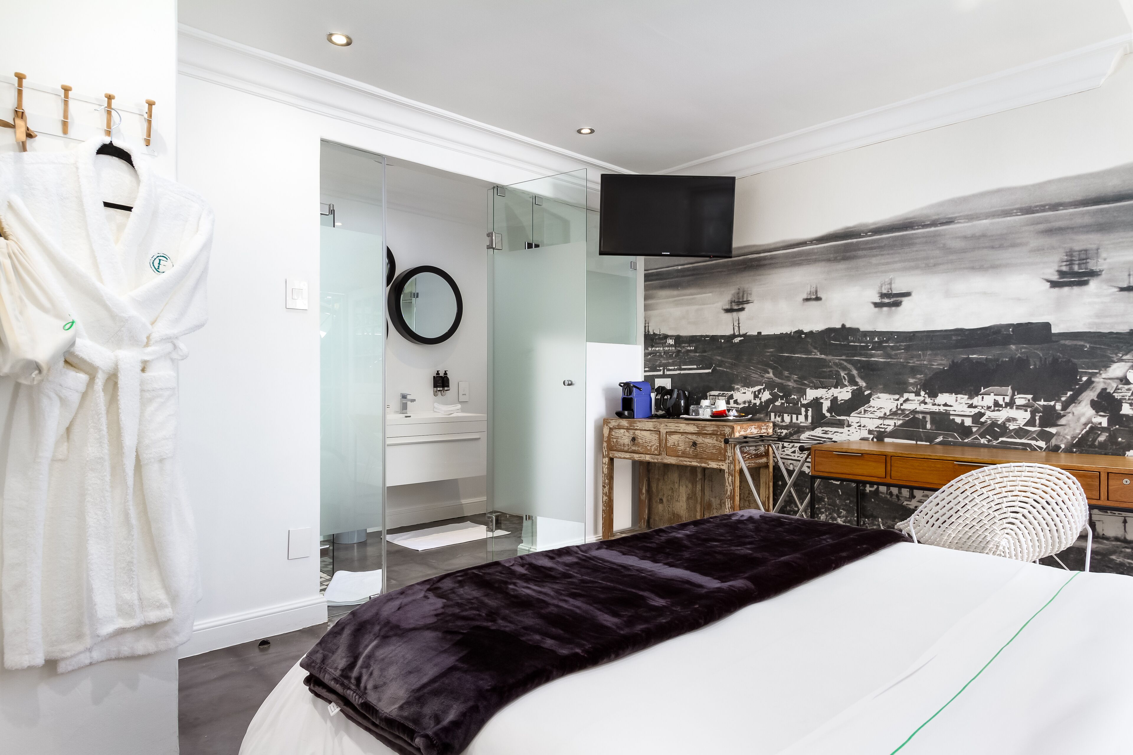 A hotel room with a bed, a desk, a chair, a TV, a robe, and a glass shower enclosure.