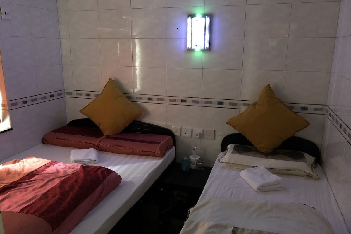 Two single beds with headboards, each with a pillow and a blanket. There is a wall socket and a green light fixture above the beds.