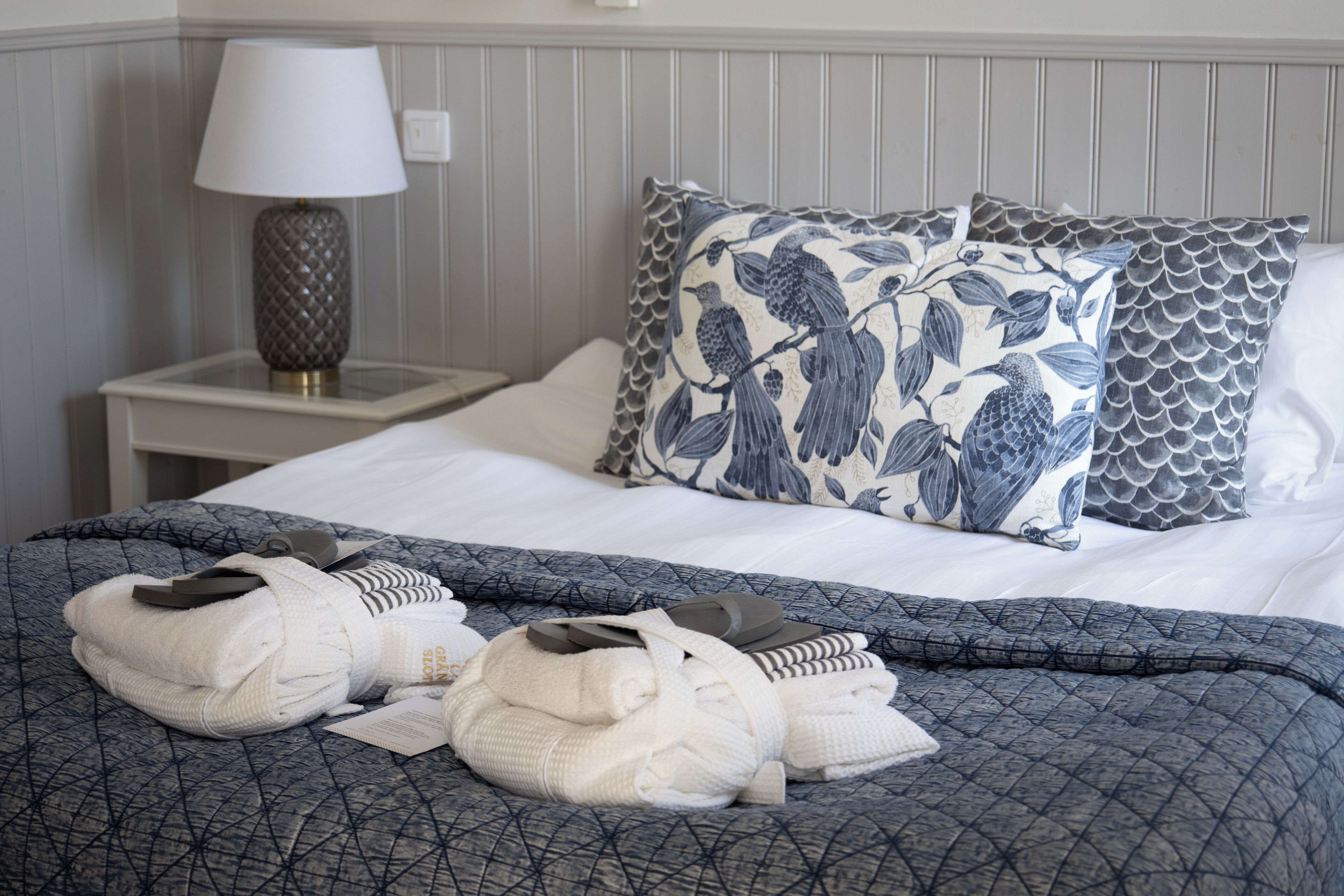 A neatly made bed with a patterned duvet, pillows with a bird design, and a lamp on a bedside table.