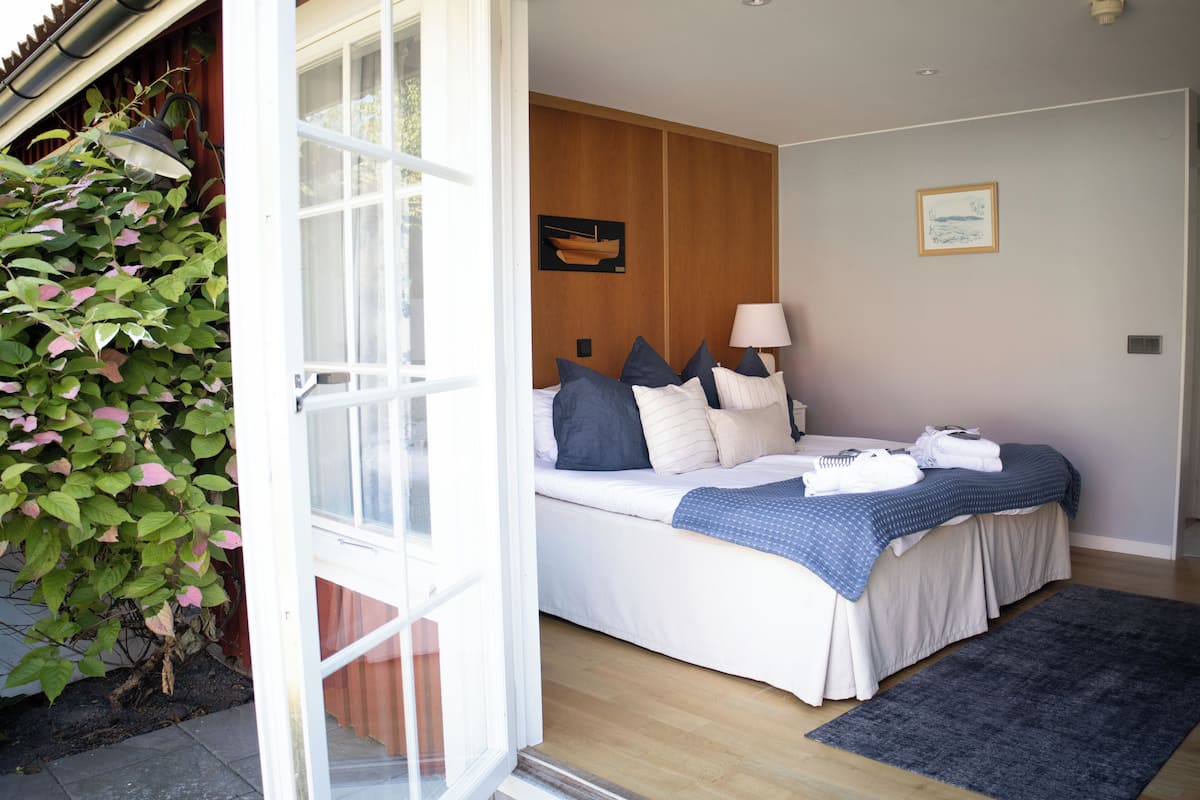 A bedroom with a bed, pillows, a bedside table, a lamp, a framed picture on the wall, and a window.