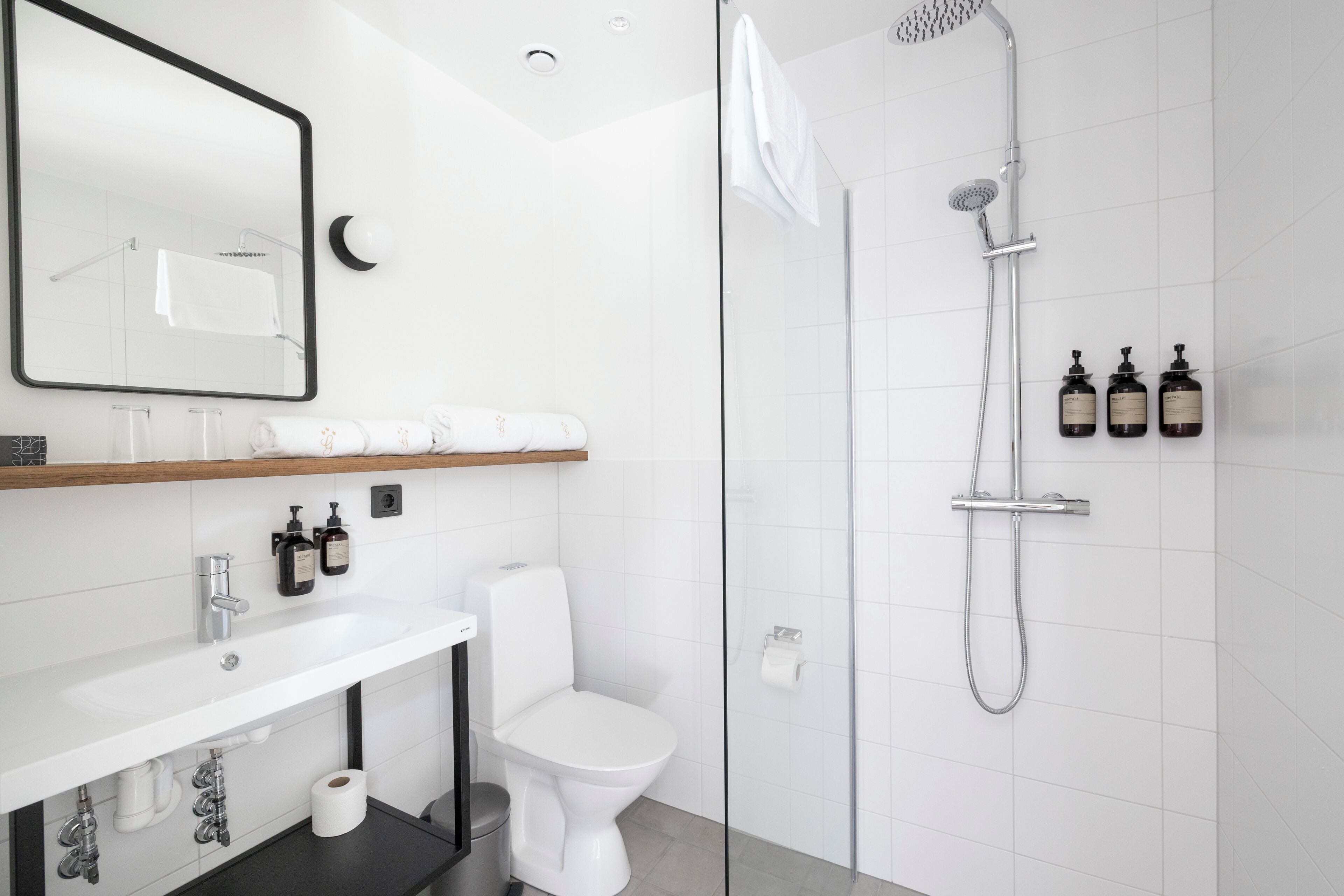 A modern bathroom with a white sink, toilet, and glass-enclosed shower. There are toiletries on the wall and a towel rack.