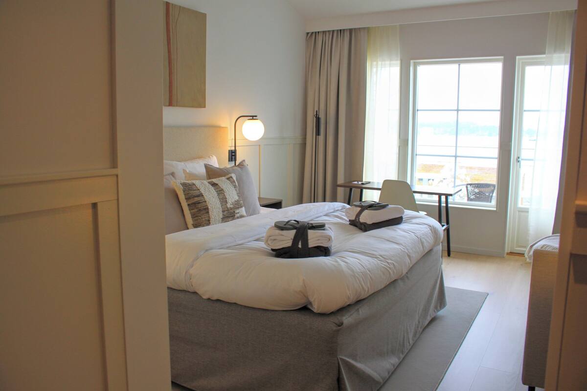 A hotel room with a large bed, a desk, a chair, a lamp, and a view of the sea through the window.
