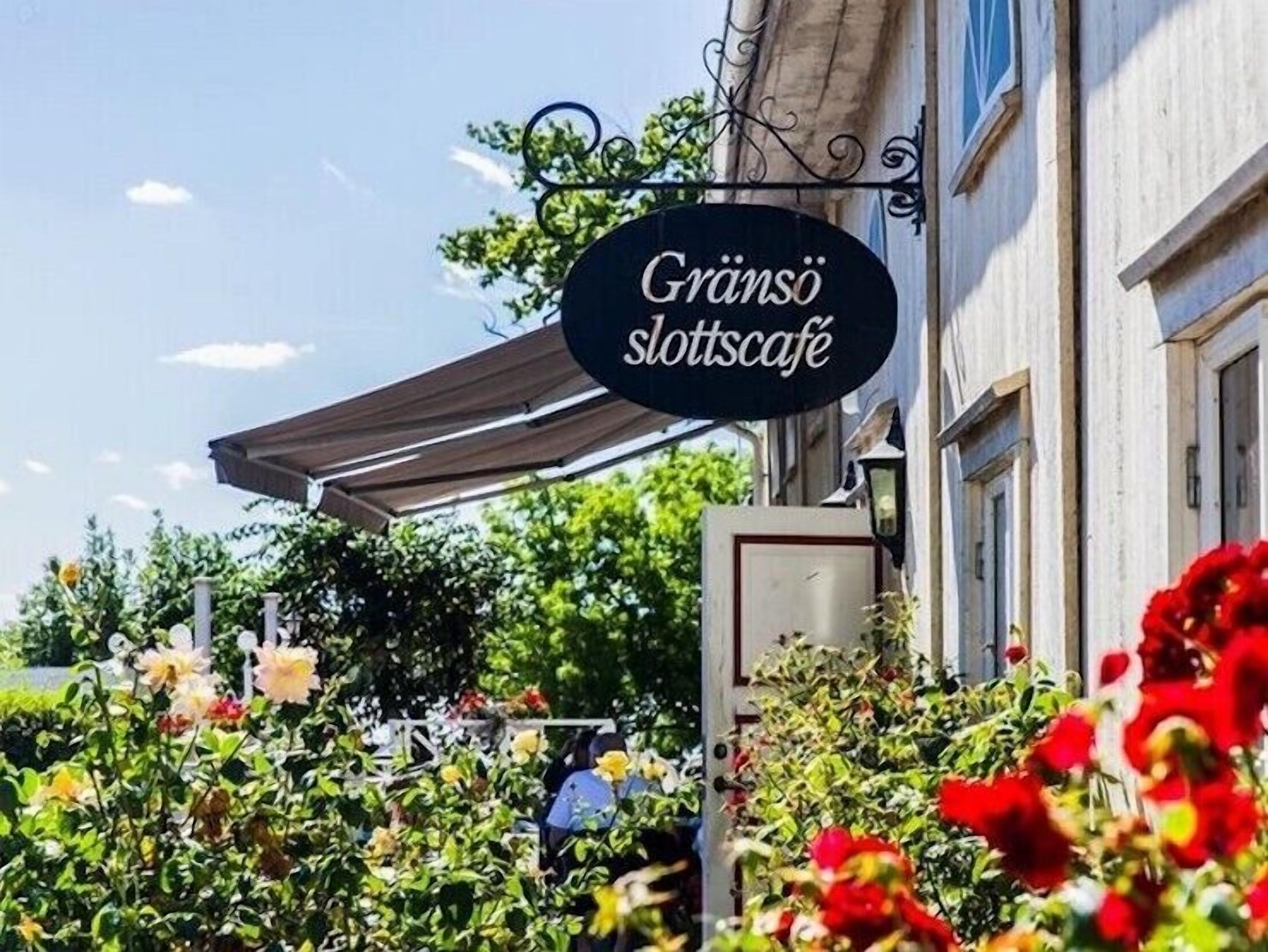 En skylt för Gräno slottscafé, med ett takförsett utomhusområde med sittplatser och en trädgård med olika sorters blommor.