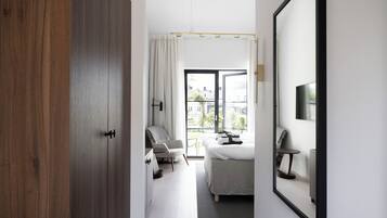 A modern hotel room with a large bed, a sitting area with a chair and table, a window with a view of buildings, and a door with a wooden panel.