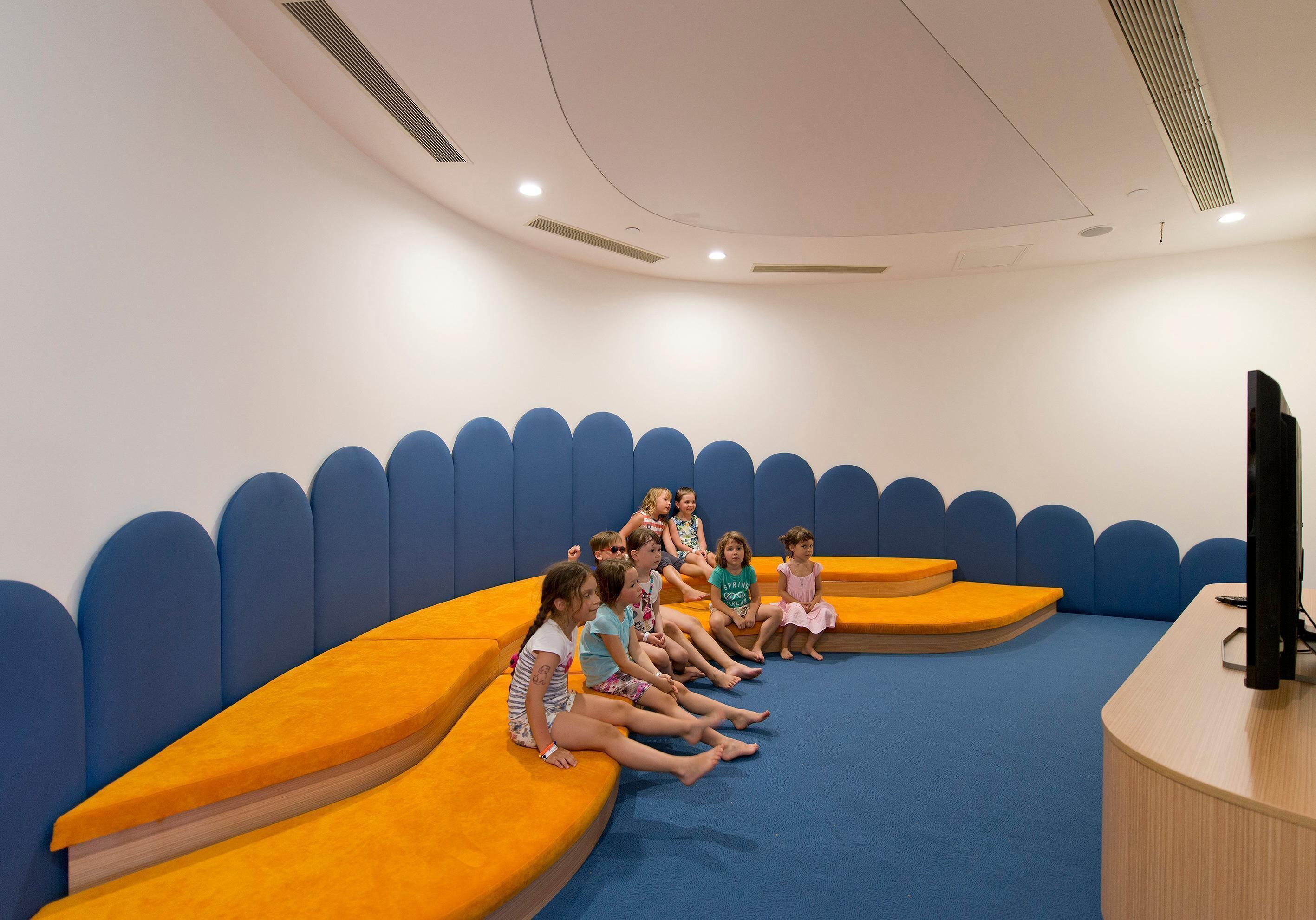 A group of children seated on orange and blue cushioned benches in a room with a curved ceiling and a flat-screen TV on a wooden table.
