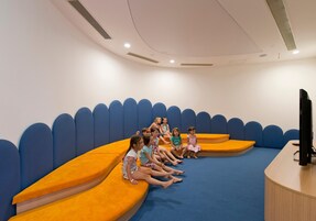 Un grupo de niños sentados en bancos acolchados de color naranja y azul en una habitación con techo curvo y un televisor de pantalla plana sobre una mesa de madera.