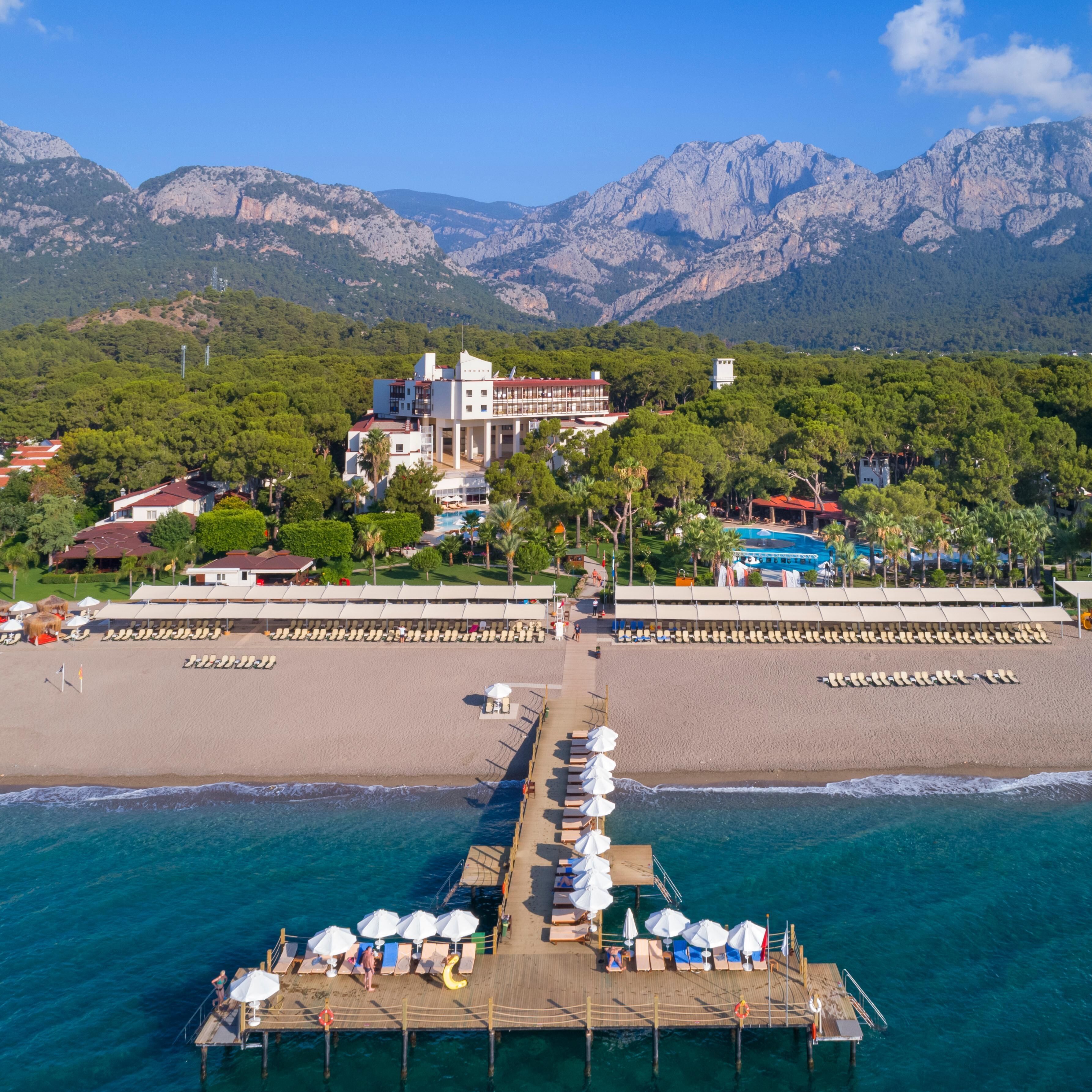 Un complexe hôtelier doté d’une plage, d’une jetée avec des chaises longues et offrant une vue sur les montagnes en arrière-plan.
