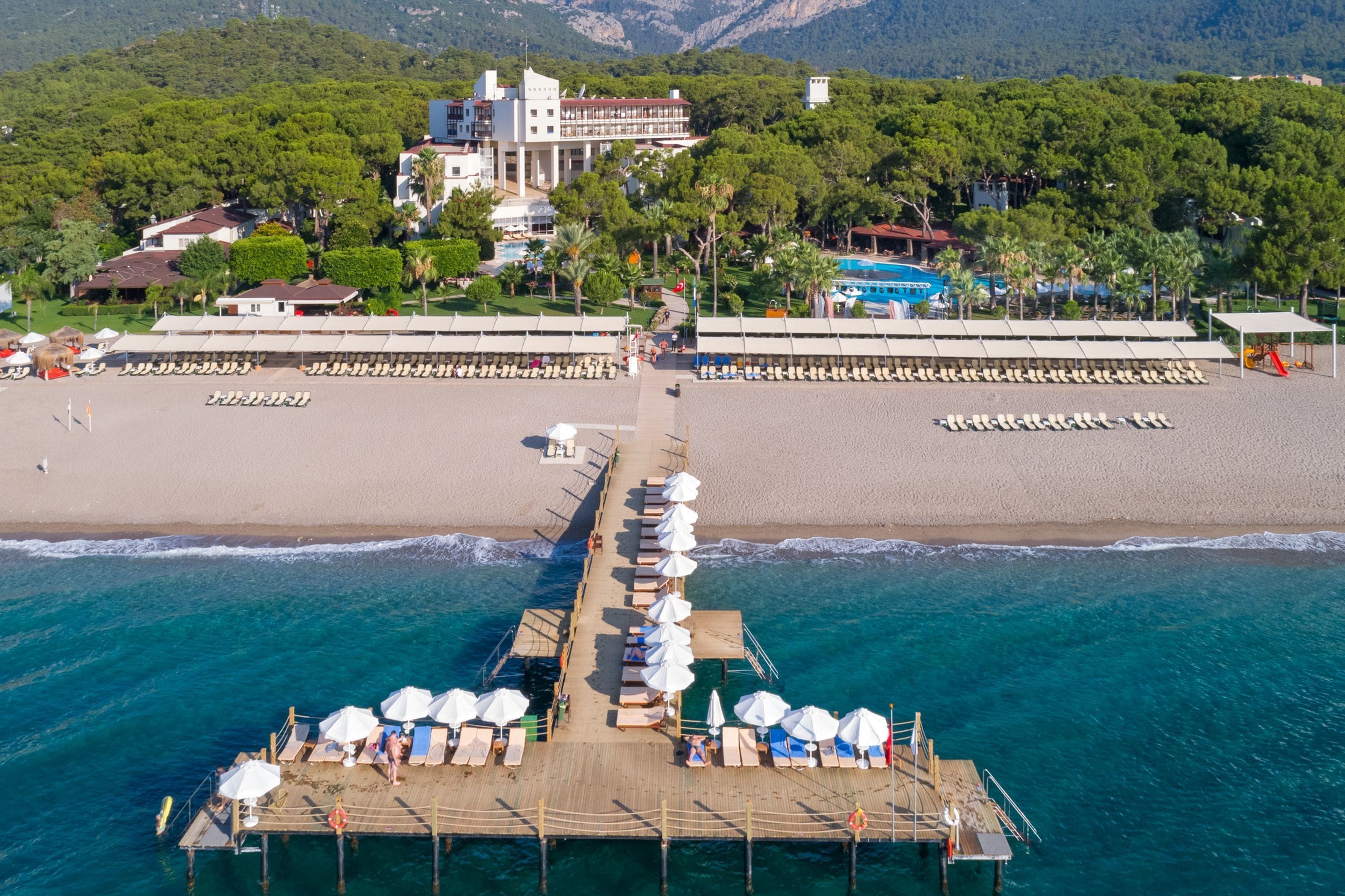 A pier with sun loungers and umbrellas, leading to a beach with a pool and a building in the background.