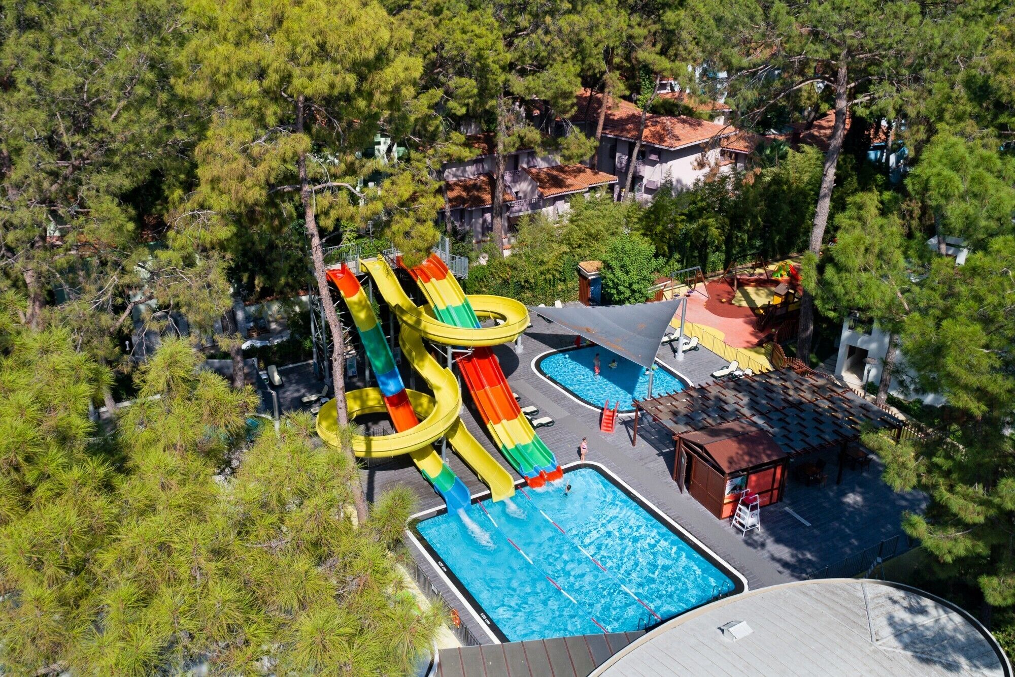 An aerial view of a resort with multiple swimming pools, a water park with slides, and surrounding pine trees.